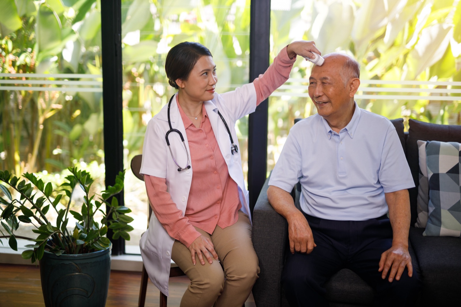 A nurse checking an older man's temperature as he sits on his sofa at home