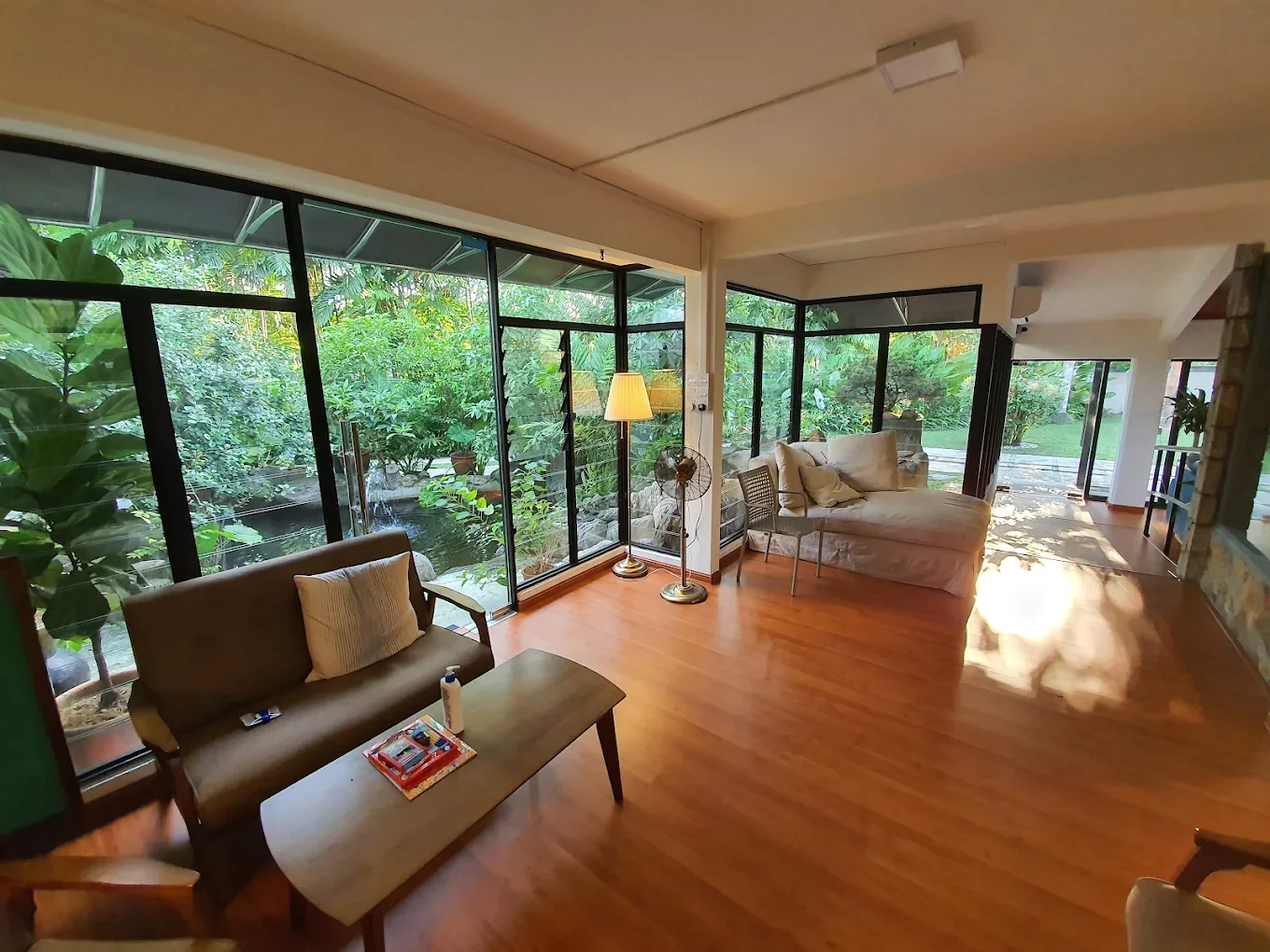 A bright lounge with a sofa, armchair and floor lamp, its glass walls looking onto a garden